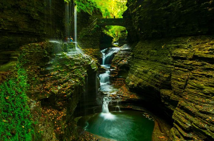 Steep rocky cliffs encase a waterfall in Watkins Glen State Park as visitors traverse the steps nearby.