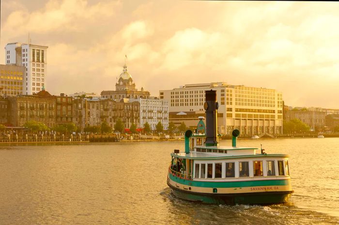 A green and white ferry approaches a historic riverside street in Savannah, Georgia.