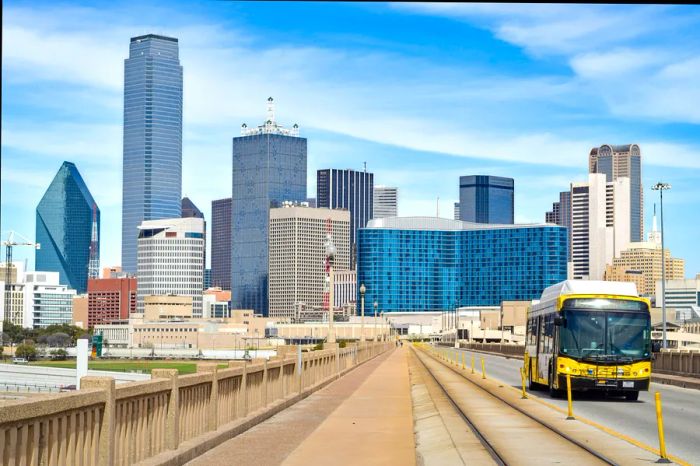 A public bus makes its way down a highway, leaving the heart of Dallas, Texas, with several skyscrapers visible in the background.