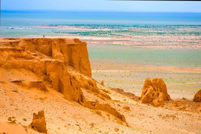 A solitary figure strides across a raised flat rock amidst a sprawling desert landscape.