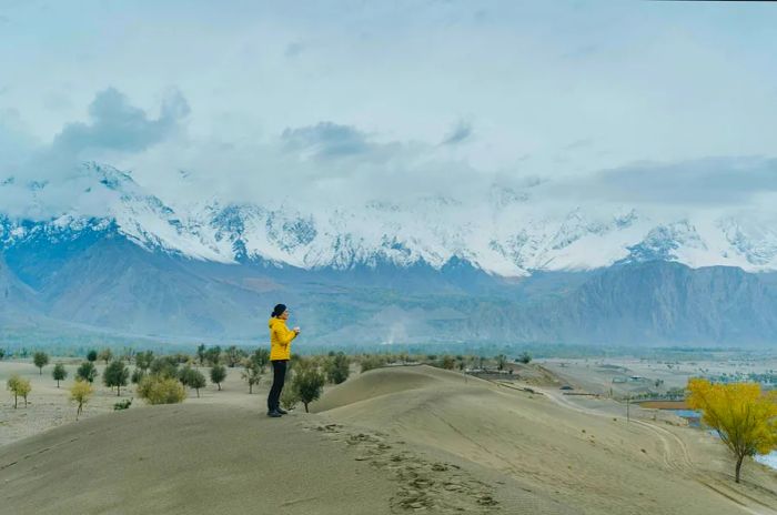 A woman stands on a sandy expanse with a backdrop of snow-capped mountains in the distance.