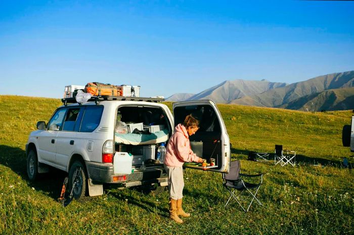 A woman stands behind a fully loaded 4WD, preparing food in a grassy meadow