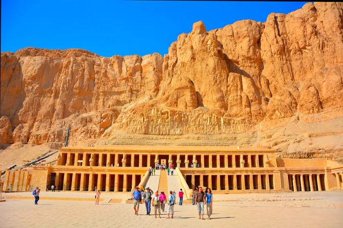 Visitors take in the magnificent facade of the Temple of Hatshepsut in Luxor, an expansive temple set against the backdrop of a rocky cliff.