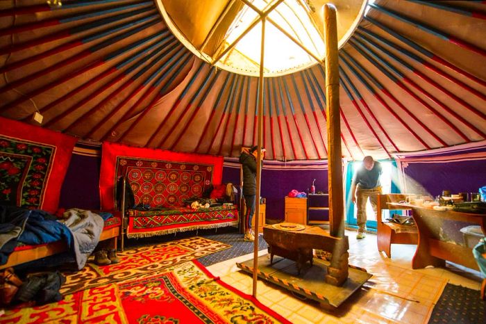 Visitors enter through the doorway of a spacious round tent featuring a central opening and adorned with vibrant rugs on both walls and floor