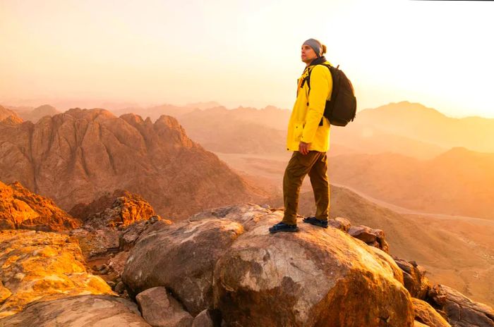 A hiker in a vivid yellow coat and a black backpack stands atop a mountain in Sinai, Egypt.