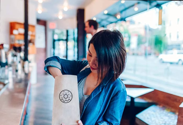 A young woman rummages through a brown paper bag featuring a doughnut outline.