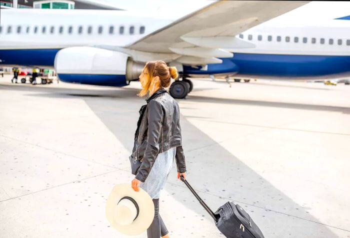 A woman in a black jacket pulls her suitcase across the tarmac while balancing a large hat.