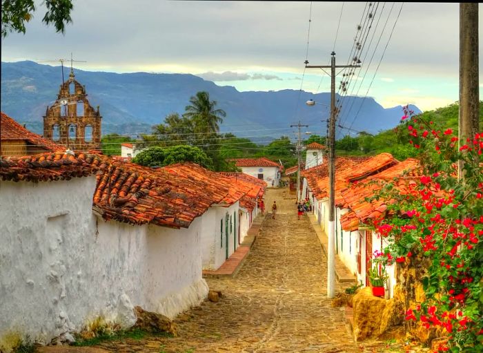 Hikers stroll along a cobblestone path through a village characterized by low-rise whitewashed buildings topped with red-tiled roofs.