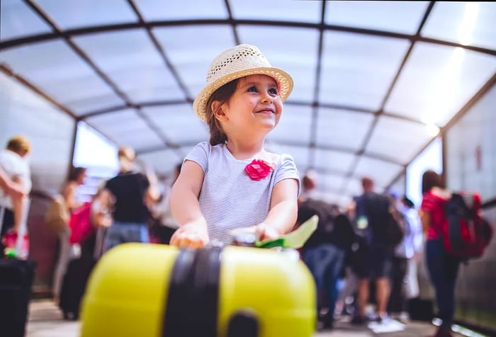 A cheerful little girl clutches a yellow suitcase, surrounded by a bustling crowd in the background.