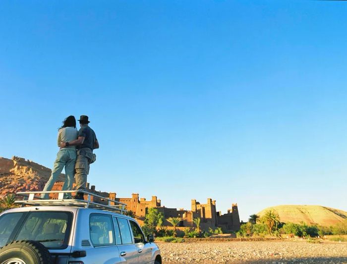 A man and woman embrace atop a four-wheel-drive vehicle, gazing at a red-stone fortress perched on a hill.