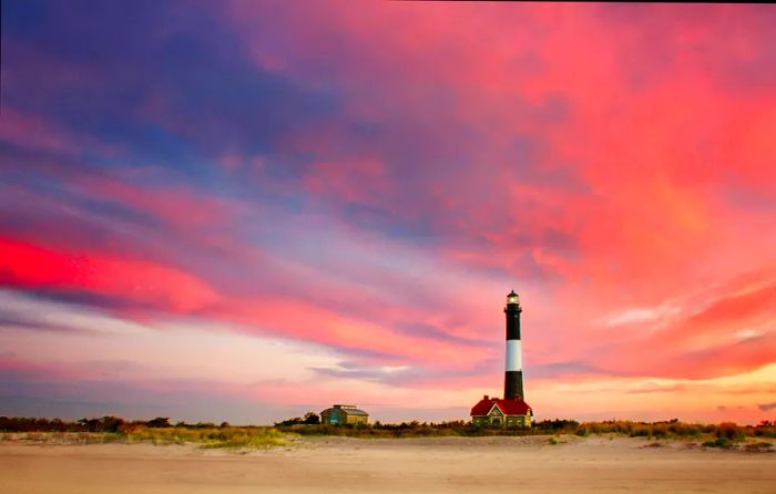 At dawn, the clouds over the Fire Island Lighthouse in Kismet, Long Island, glow in shades of pink and magenta against a backdrop of blue sky, viewed from the beach by the lighthouse.