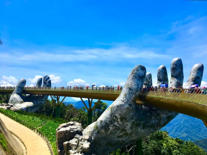 Individuals stroll across a bridge supported by two enormous stone hands in a mountain resort.