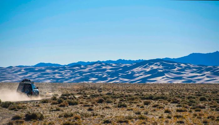 A 4WD vehicle travels along a dusty path leading to sand dunes in a desert