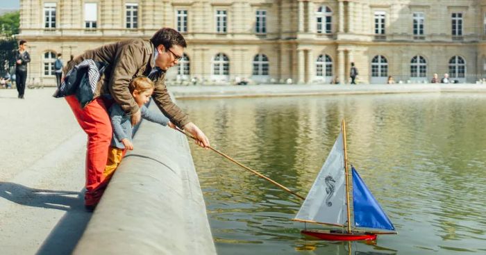 A father and son launch a small sailboat onto a pond in front of a grand palace.