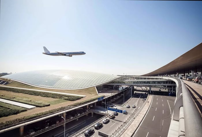 An airplane soaring above a modern airport with a curved glass roof.