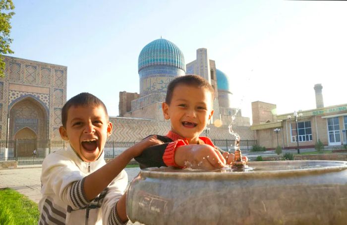 Two boys play joyfully in a small water fountain located in front of a beautifully tiled mosque.