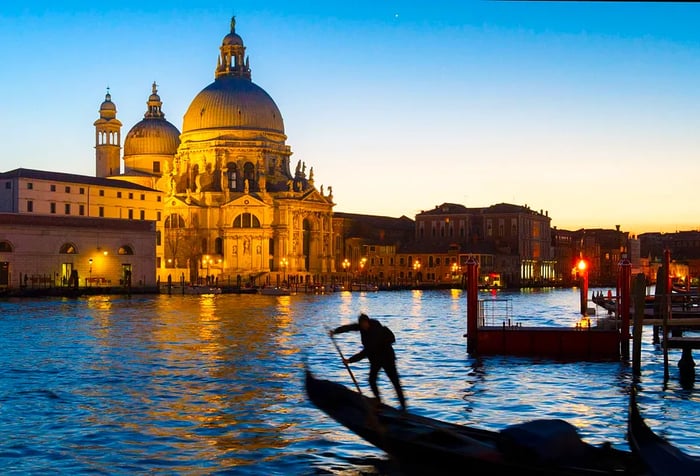 A gondolier stands silhouetted against the Grand Canal at Punta della Dogana, with the beautifully lit St. Mary of Health (Santa Maria della Salute) Basilica in the background at dusk. Venice, Veneto, Italy.