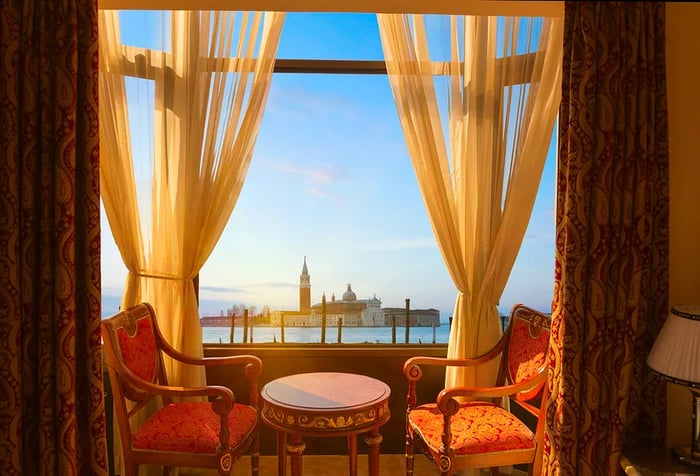 A picturesque view of the Venice lagoon from a luxurious hotel, featuring chairs and curtains in the foreground.