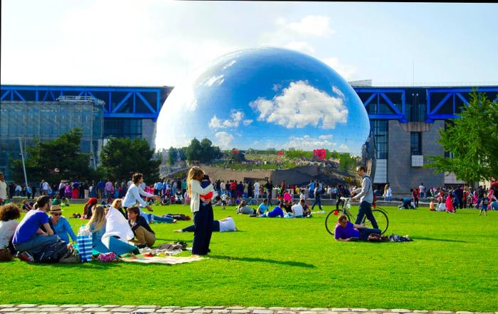 A crowd gathers in a Paris park near a large silver sphere, part of the Cité des Sciences.