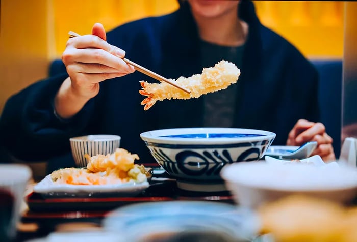 A person uses chopsticks to pick up a piece of tempura, surrounded by an array of delicious Japanese dishes on the table.