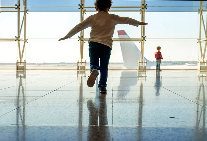 A boy standing on one foot while peering out a glass window at an airplane at the airport.