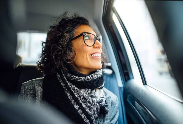 A mature woman smiles while seated in the backseat of a car, gazing out the window. A mid-aged female enjoying a car ride.