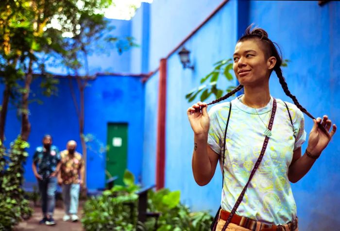 A young woman in a yard of a blue-walled building, holding her braided hair, while two men walk along a path behind her.