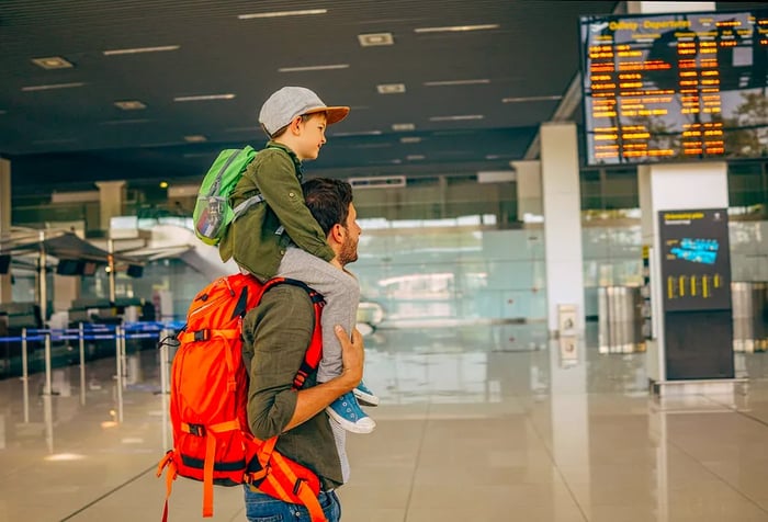 A man with an orange bag is carrying a child on his shoulders while at the airport.