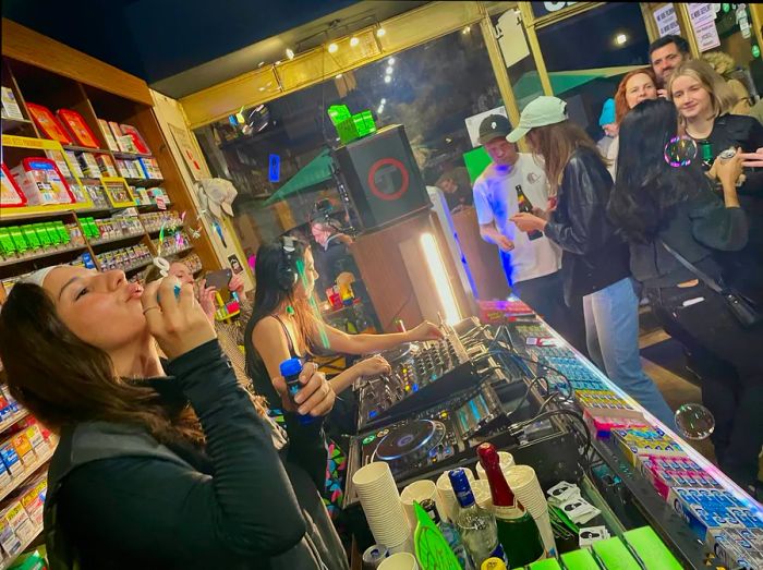 Two women DJing in a Berlin <em>Späti</em> while one blows bubbles