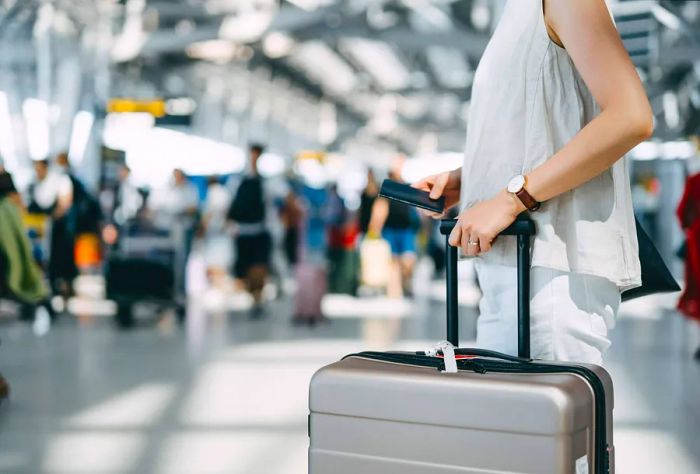 In the lively international airport terminal, a young woman strides confidently with her passport in hand, dragging her suitcase alongside her as the bustling environment fades into the background.