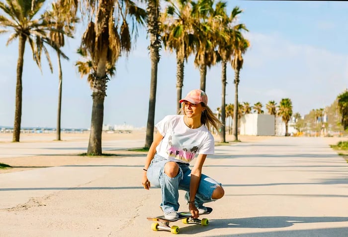 A woman skateboarding along a paved path by the beach, flanked by tall trees.