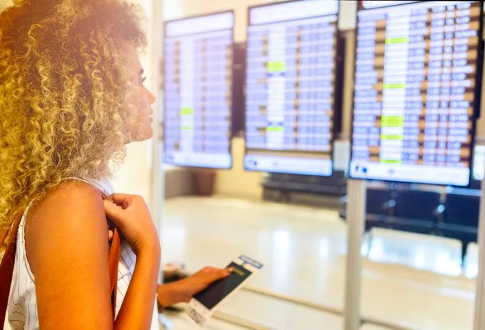 A woman with curly hair examines the airport flight information screens while clutching her passport and boarding pass.