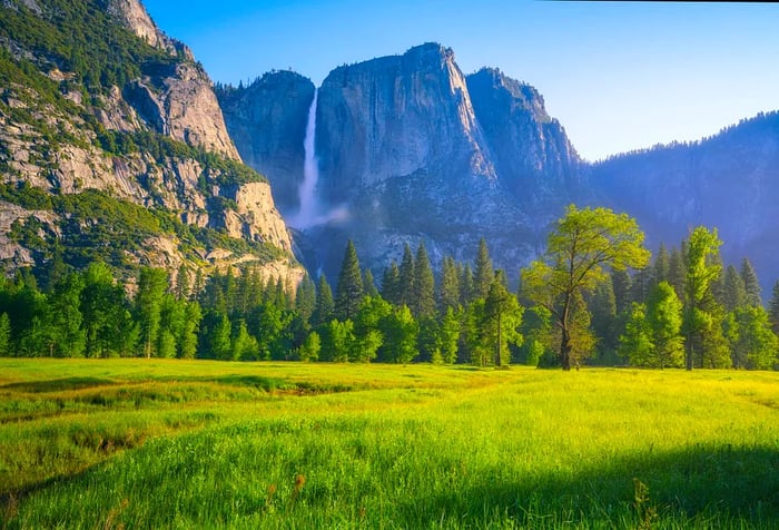 A lush valley encircled by towering green trees beneath a jagged mountain range featuring a waterfall.