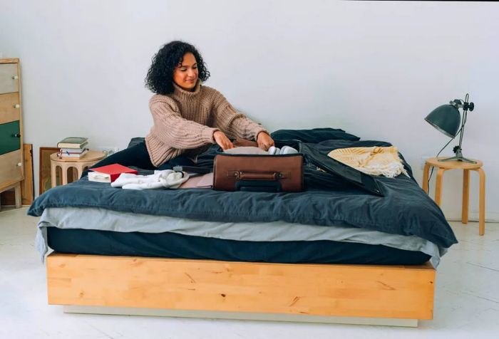 A woman with curly hair is arranging her clothes inside a brown suitcase on the bed.