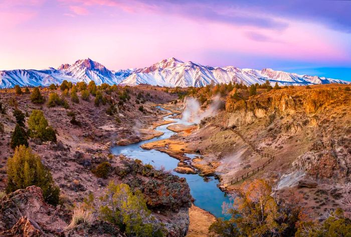 A creek winds through arid land, framed by a majestic snow-covered mountain range.