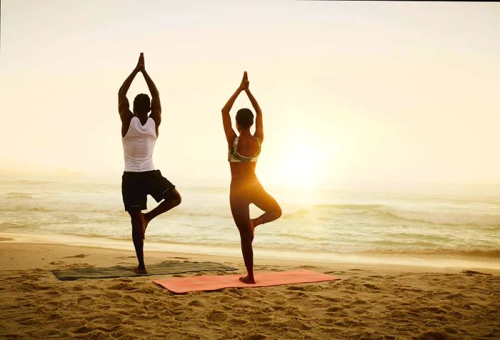 A man and woman performing yoga poses with one leg extended on a beach.