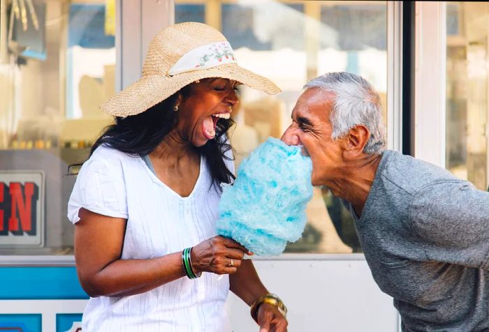 A woman with her mouth wide open enjoying a stick of cotton candy, while a man takes a hearty bite.