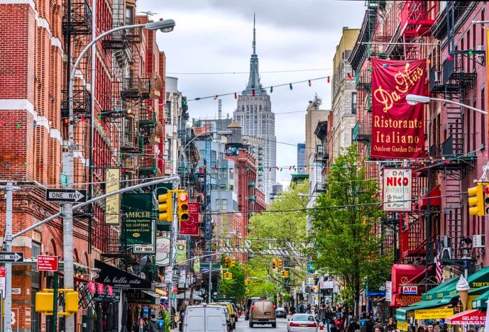 A bustling street scene in Little Italy, showcasing the Empire State Building with cars passing by and buildings lining the road.