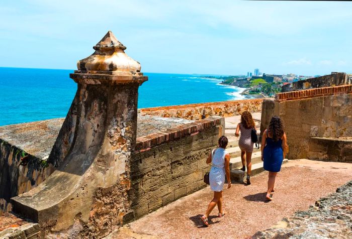 Three young women strolling along a path of an ancient structure perched on a cliff with ocean views.