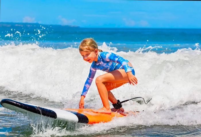 A cheerful young girl with blonde hair balances on a surfboard as waves crash behind her.