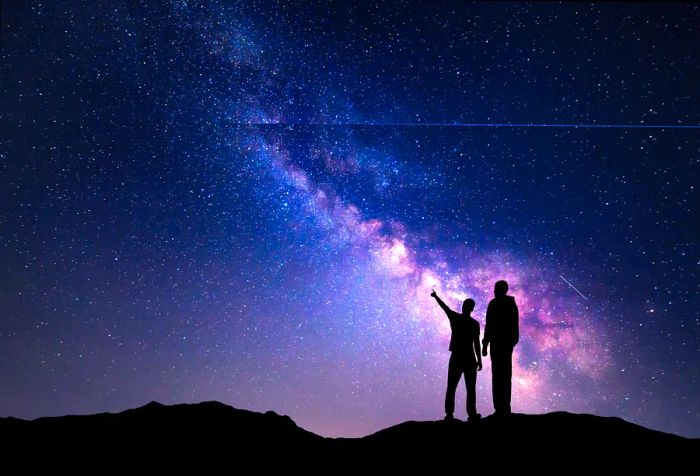 A silhouette of a family atop a mountain, with a father and son pointing at the starry night sky filled with the Milky Way. A beautiful cosmic landscape that encapsulates the wonder of the universe—perfect for travel enthusiasts.