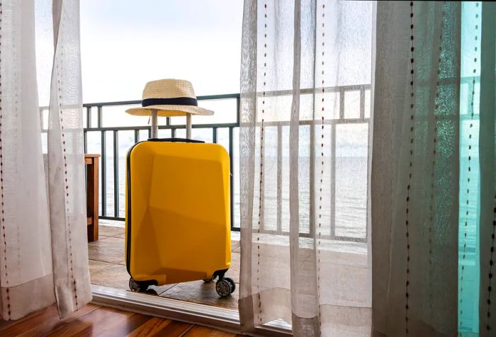 A vibrant yellow suitcase topped with a hat, positioned on a patio behind glass doors.