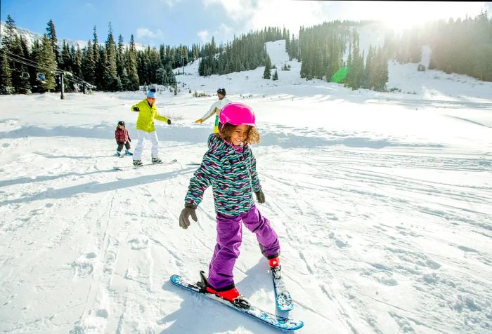 A little girl carefully skis with others following her in a snowy forest.