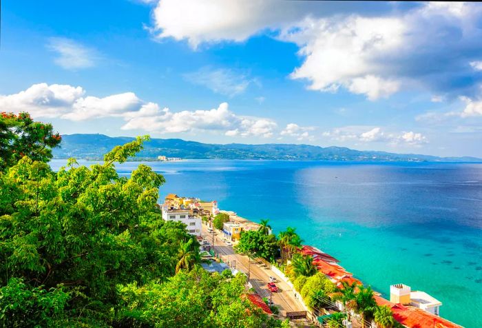 Aerial view showcasing a lush coastal town that overlooks the serene turquoise sea, framed by a mountain range under a partly cloudy blue sky.