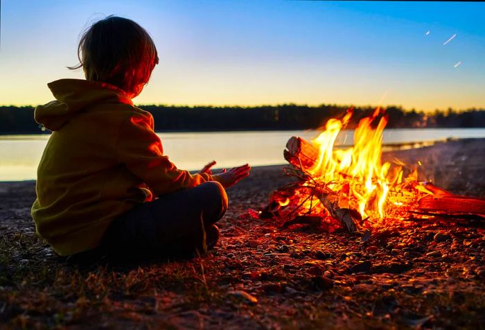 A child wearing a hooded jacket sits cross-legged before a bonfire, extending open hands towards the warmth.