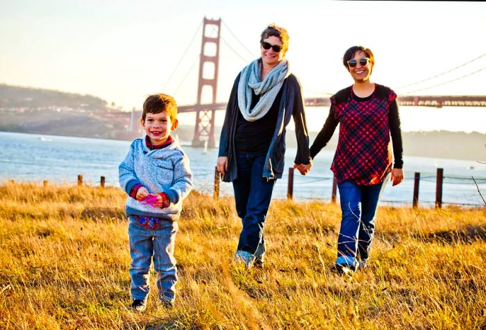 A family of three poses in a grassy field with the San Francisco bridge visible in the background.