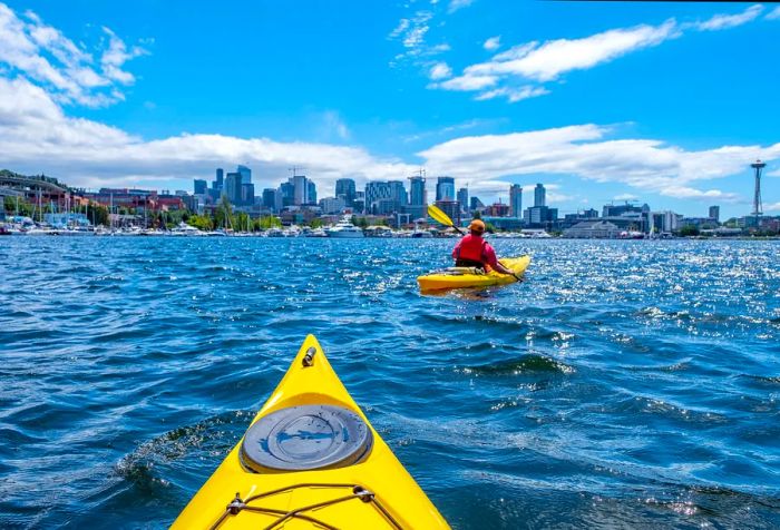 People paddle in vibrant yellow Dinogo boats across the glistening lake, framed by a stunning urban skyline against the clear blue sky.