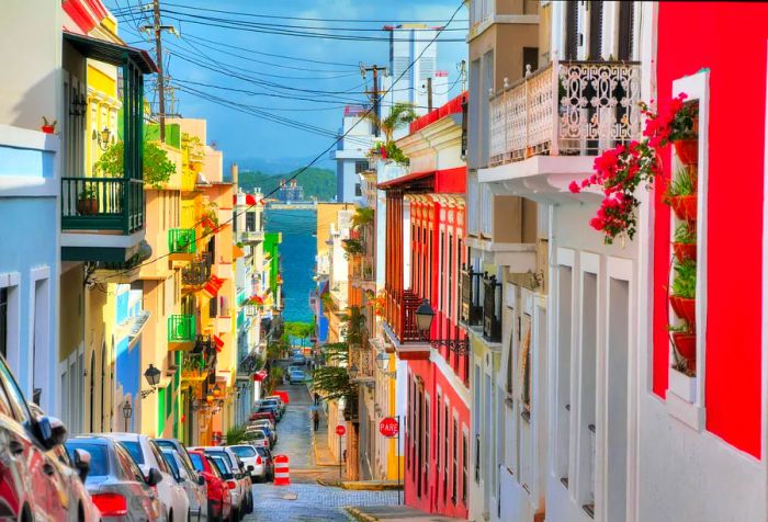 A narrow street lined with parked cars on one side and vibrant buildings on the other.