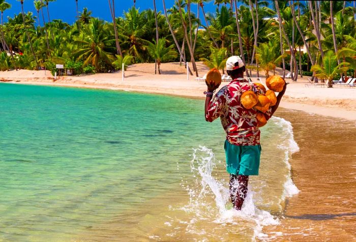 A man wearing a printed shirt and white cap strolls through the shallow waters of a beach, carrying coconuts toward a cluster of palm trees.