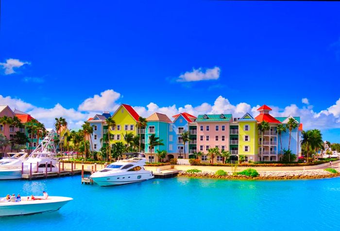 Vibrant houses line the shore, with yachts anchored against the backdrop of the turquoise sea and a partly cloudy blue sky.